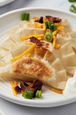 Lightly-fried pierogies, fanned on a white plate and garnished with bacon, green onions, and cheese.